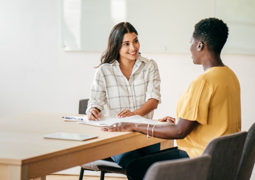 A property manager with long, dark hair speaks to a landlord with short hair over a desk with paperwork.